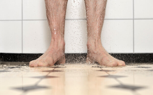 Mann steht unter der Dusche und betreibt Intimpflege, um einer Posthitis vorzubeugen, dabei sieht man nur seine Füße und Waden.