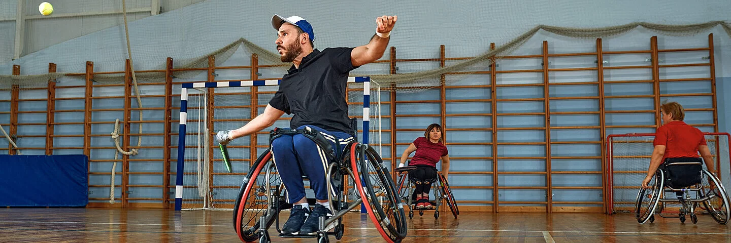 Mann im Rollstuhl spielt Tennis in einer Turnhalle.