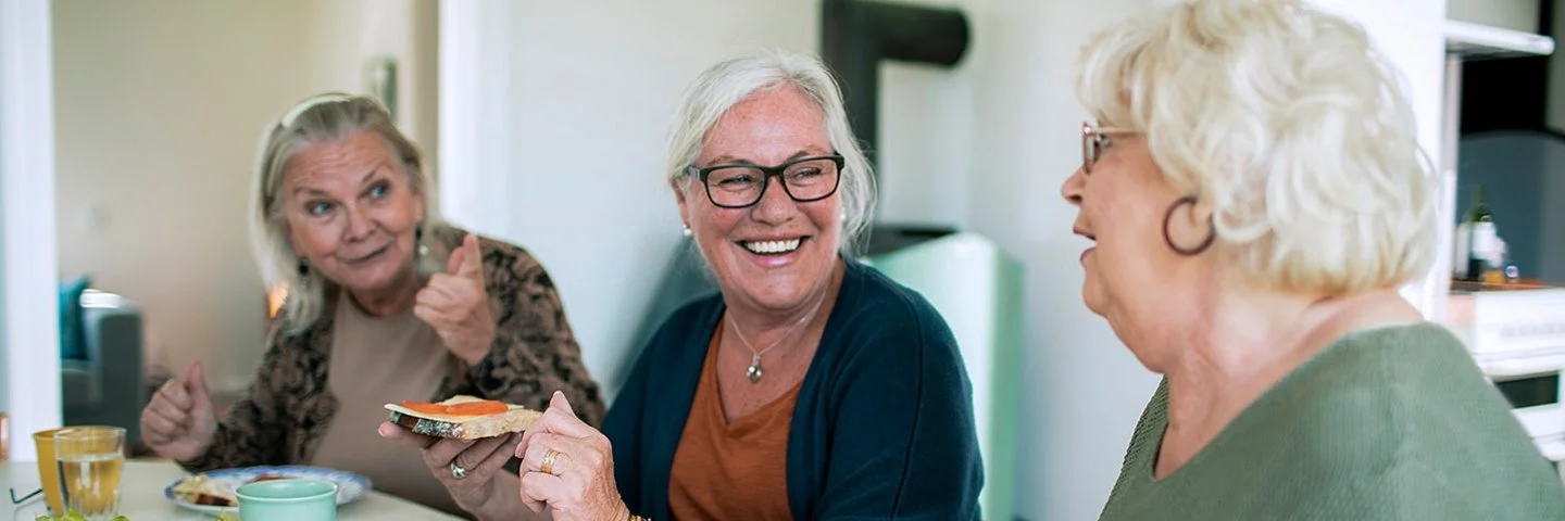 Drei ältere Frauen sitzen zusammen in der gemeinsamen Wohnung am Frühstückstisch. Sie frühstücken und unterhalten sich dabei lachend. Auf dem Tisch stehen, Geschirr, Brot, Brotbelag und aufgeschnittenes Gemüse. Im Hintergrund ist ein Teil der Küche zu sehen.