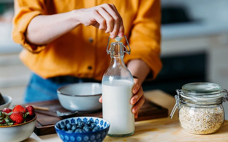 Auf einer Küchenarbeitsplatte stehen eine Schale mit Erdbeeren, eine Schale mit Blaubeeren und ein Einweckglas mit Haferflocken. Eine Person, deren Kopf nicht im Bild ist, verschließt eine Milchflasche.