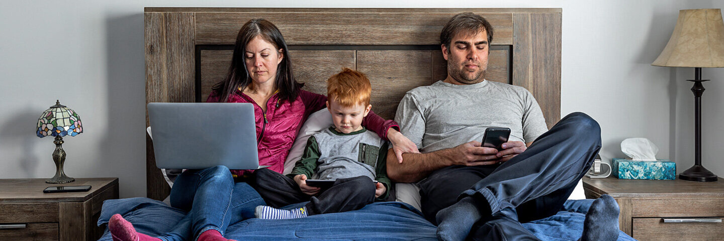 Vater, Mutter und kleiner Sohn sitzen im Bett – alle sind durch digitale Endgeräte abgelenkt.