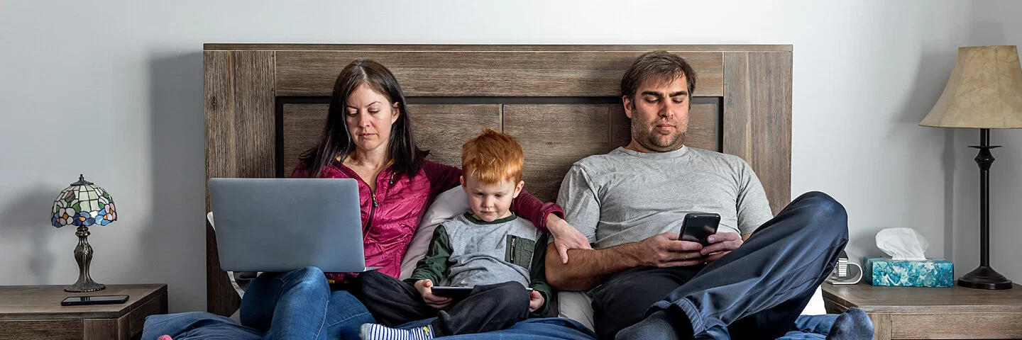 Vater, Mutter und kleiner Sohn sitzen im Bett – alle sind durch digitale Endgeräte abgelenkt.