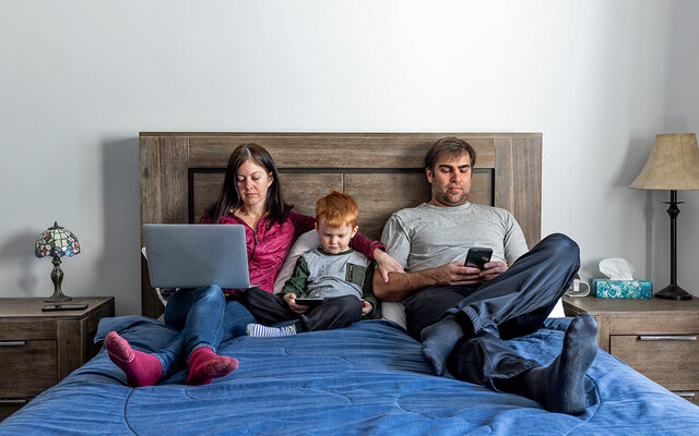 Vater, Mutter und kleiner Sohn sitzen im Bett – alle sind durch digitale Endgeräte abgelenkt.