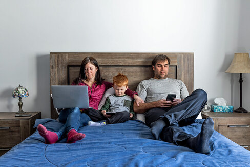 Vater, Mutter und kleiner Sohn sitzen im Bett – alle sind durch digitale Endgeräte abgelenkt.