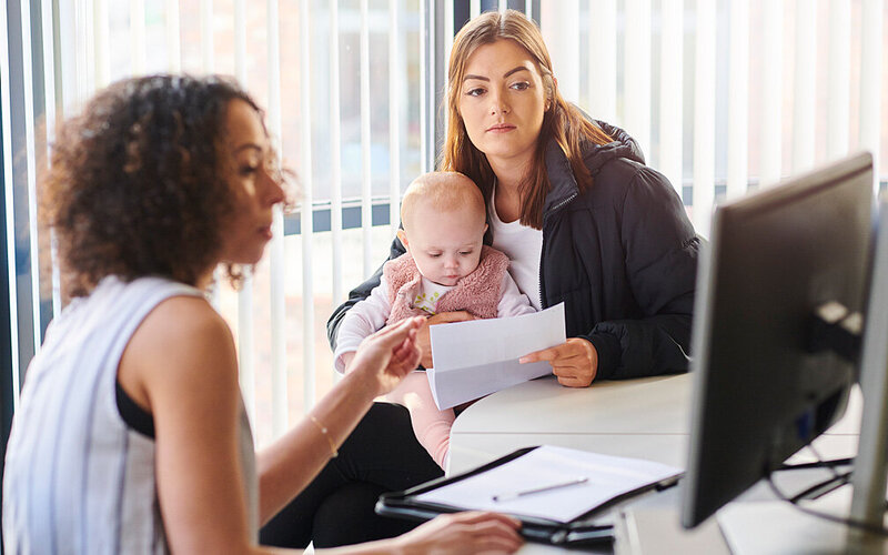 Eine junge Mutter mit Baby auf dem Schoß sitzt im Büro einer Sachbearbeiterin und bespricht Unterstützungsangebote.