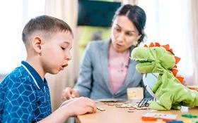 Ein Junge übt gemeinsam mit einer Therapeutin das Sprechen. Eine grüne Dinosaurier-Handpuppe unterstützt die Übung.