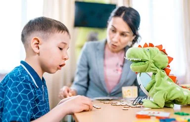 Ein Junge übt gemeinsam mit einer Therapeutin das Sprechen. Eine grüne Dinosaurier-Handpuppe unterstützt die Übung.