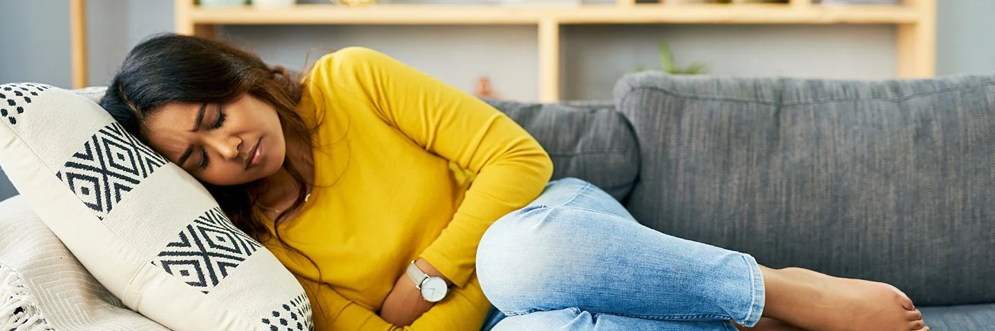 Eine Frau liegt gekrümmt auf dem Sofa und hält sich den Bauch. 