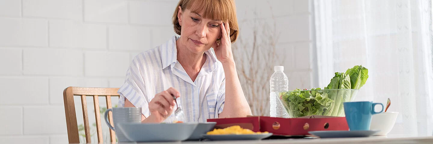 Eine Frau mit Reizmagen sitzt vor ihrem Essen, mit einer Hand hält sie eine Gabel, mit der anderen stützt sie ihren Kopf, auf dem Tisch stehen eine Schüssel Salat und eine Flasche Wasser.