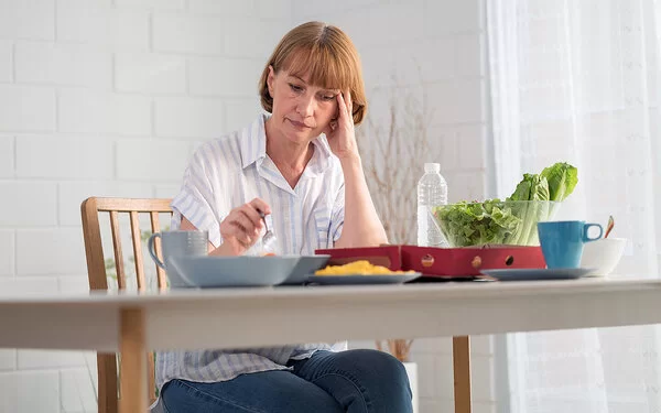 Eine Frau mit Reizmagen sitzt vor ihrem Essen, mit einer Hand hält sie eine Gabel, mit der anderen stützt sie ihren Kopf, auf dem Tisch stehen eine Schüssel Salat und eine Flasche Wasser.