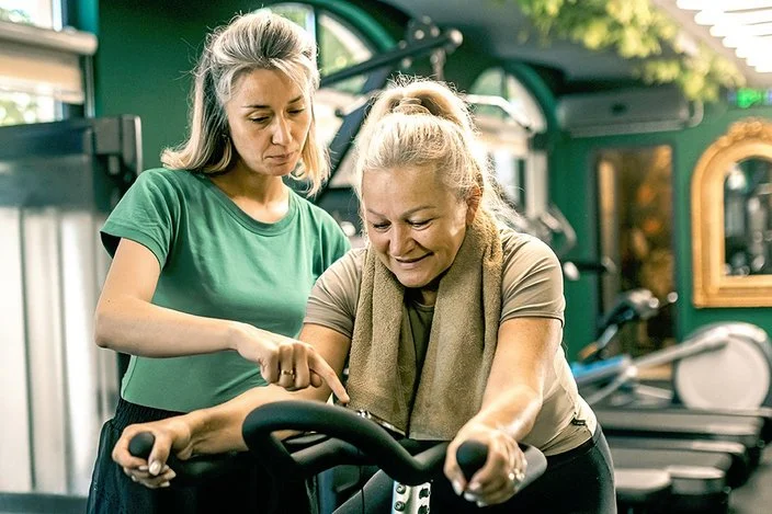 In einem Fitnessstudio sitzt eine ältere Frau auf einem Heimtrainer. Eine jüngere Frau mit grauen Haaren und grünem T-Shirt steht daneben und zeigt auf das Display des Fahrrads.