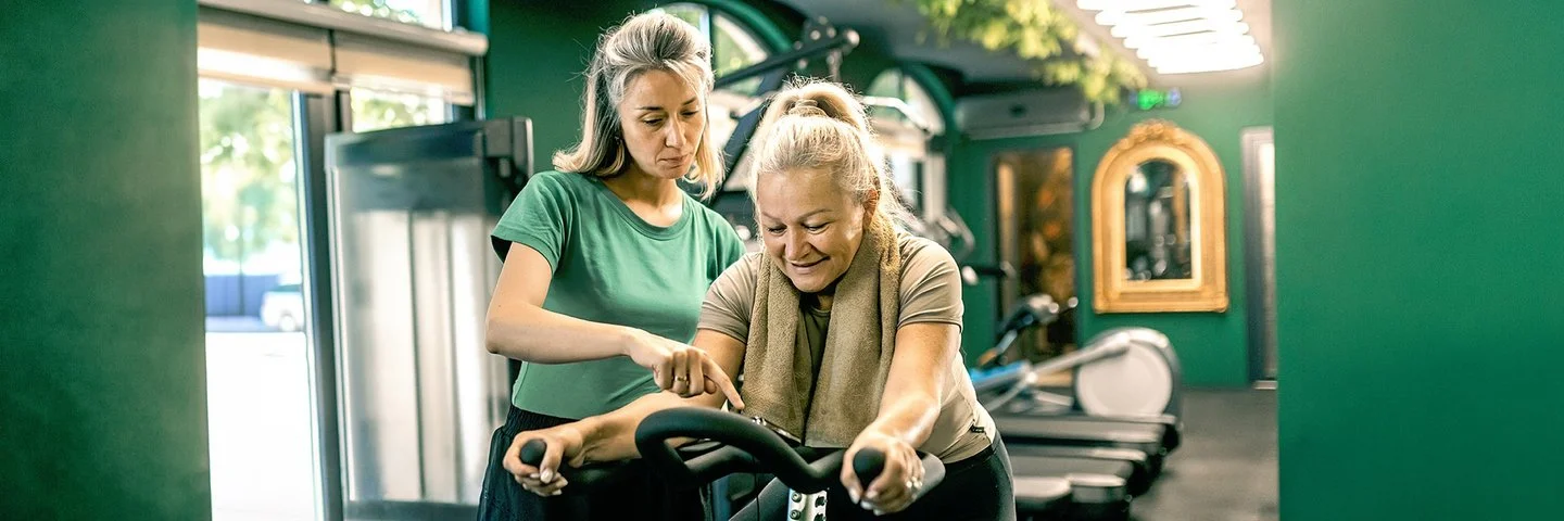 In einem Fitnessstudio sitzt eine ältere Frau auf einem Heimtrainer. Eine jüngere Frau mit grauen Haaren und grünem T-Shirt steht daneben und zeigt auf das Display des Fahrrads.