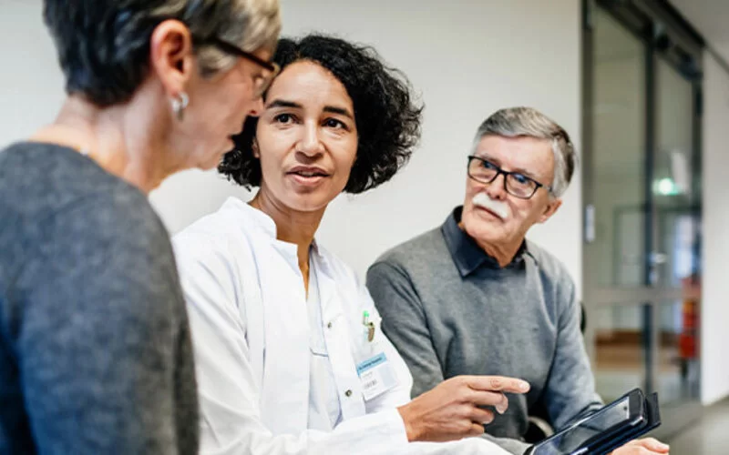 Eine junge Ärztin sitzt neben zwei älteren Personen und unterhält sich mit ihnen.