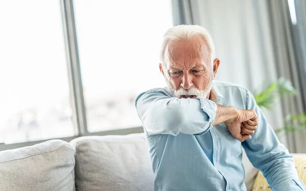 Ein älterer Mann sitzt auf einem Sofa und hustet in seine rechte Armbeuge.