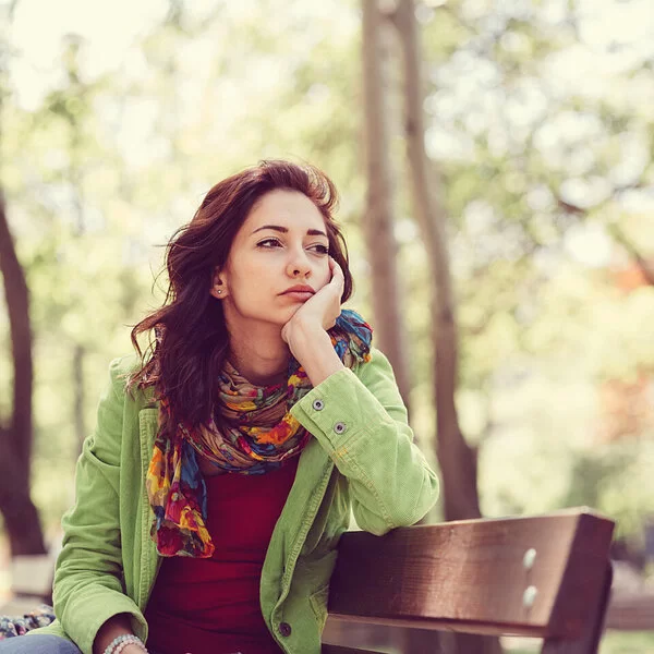 Eine junge Frau sitzt allein auf einer Parkbank; die rechte Wange in die Handfläche gestützt, schaut sie traurig in die Ferne.