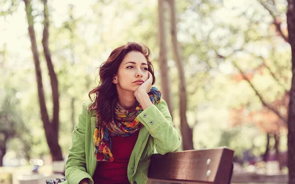 Eine junge Frau sitzt allein auf einer Parkbank; die rechte Wange in die Handfläche gestützt, schaut sie traurig in die Ferne.