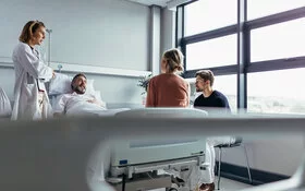 In einem Krankenzimmer redet eine Ärztin mit zwei Angehörigen und dem im Bett liegenden Patienten.