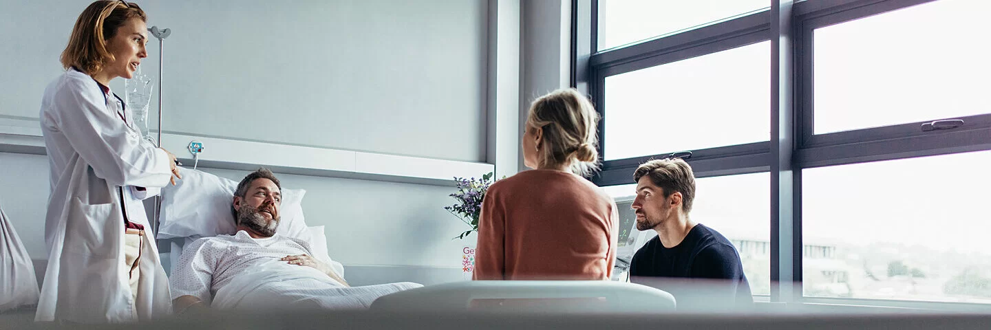 In einem Krankenzimmer redet eine Ärztin mit zwei Angehörigen und dem im Bett liegenden Patienten.