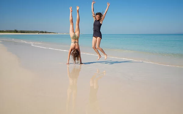 Zwei junge Frauen am Strand, eine macht einen Handstand, die andere springt mit erhobenen Händen in die Höhe.