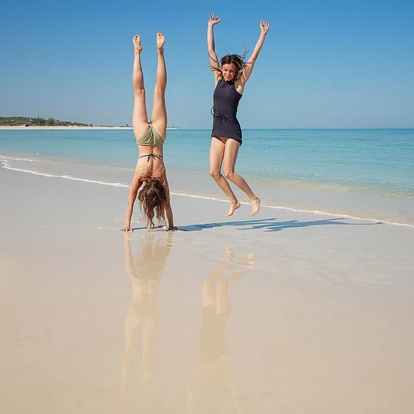 Zwei junge Frauen am Strand, eine macht einen Handstand, die andere springt mit erhobenen Händen in die Höhe.