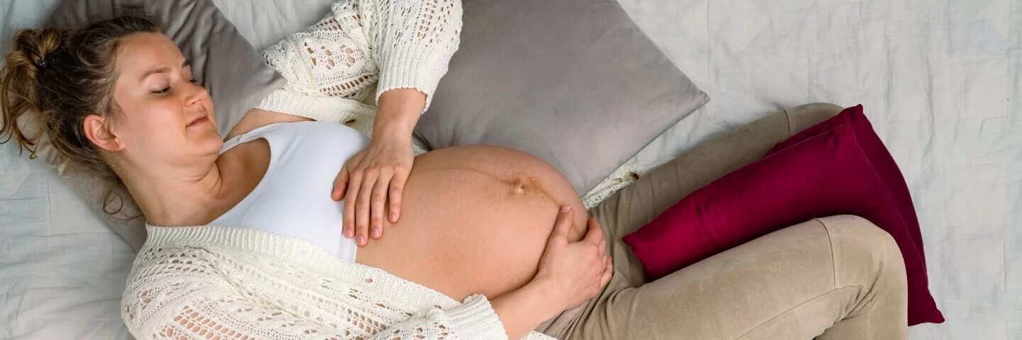 Eine schwangere Frau liegt auf einem Bett und hält ihren Bauch, auf dem eine Linea negra erkennbar ist.