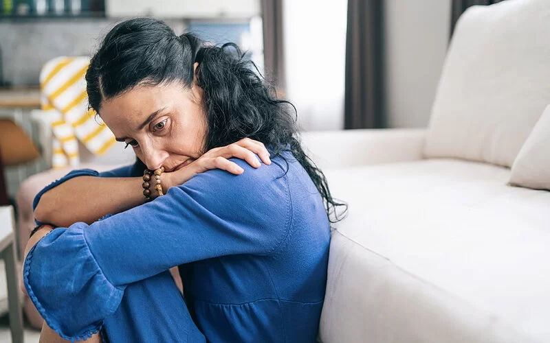 Eine Frau, die unter Agoraphobie leidet und in sich gekehrt auf dem Boden an ein Sofa gelehnt sitzt.