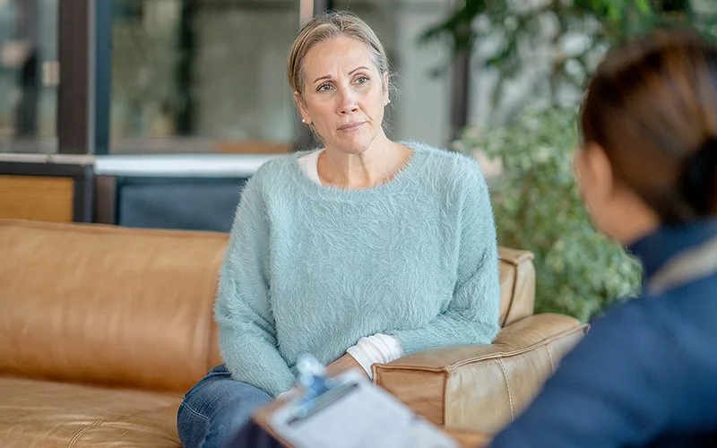 Eine Frau in einem flauschigen hellblauen Pullover sitzt auf einem braunen Ledersofa und blickt aufmerksam auf eine Person, die im Vordergrund ein Klemmbrett hält.