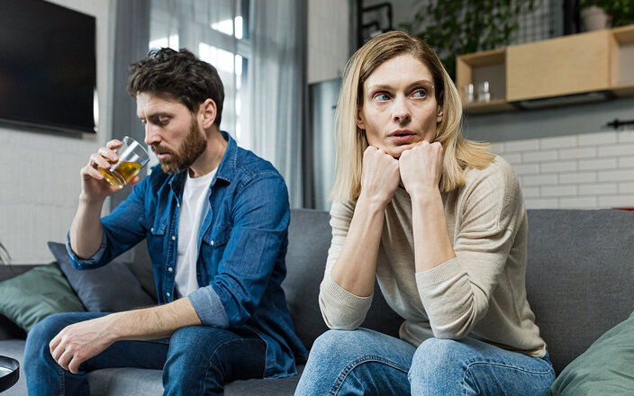 Co-Abhängigkeit: Ein alkoholkranker Mann trinkt auf dem Sofa neben seiner müde und blass aussehenden Partnerin, die zur Seite wegschaut.