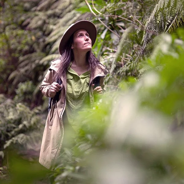 Eine Frau, die langärmelige Kleidung und einen breitkrempigen Hut trägt, wandert durch ein dichtes grünes Dickicht. Die Vegetation besteht hauptsächlich aus großen Farnen.