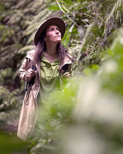 Eine Frau, die langärmelige Kleidung und einen breitkrempigen Hut trägt, wandert durch ein dichtes grünes Dickicht. Die Vegetation besteht hauptsächlich aus großen Farnen.