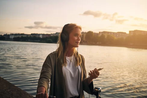 Eine junge Frau steht an einem Flussufer und hört mit Kopfhörern einen Podcast.