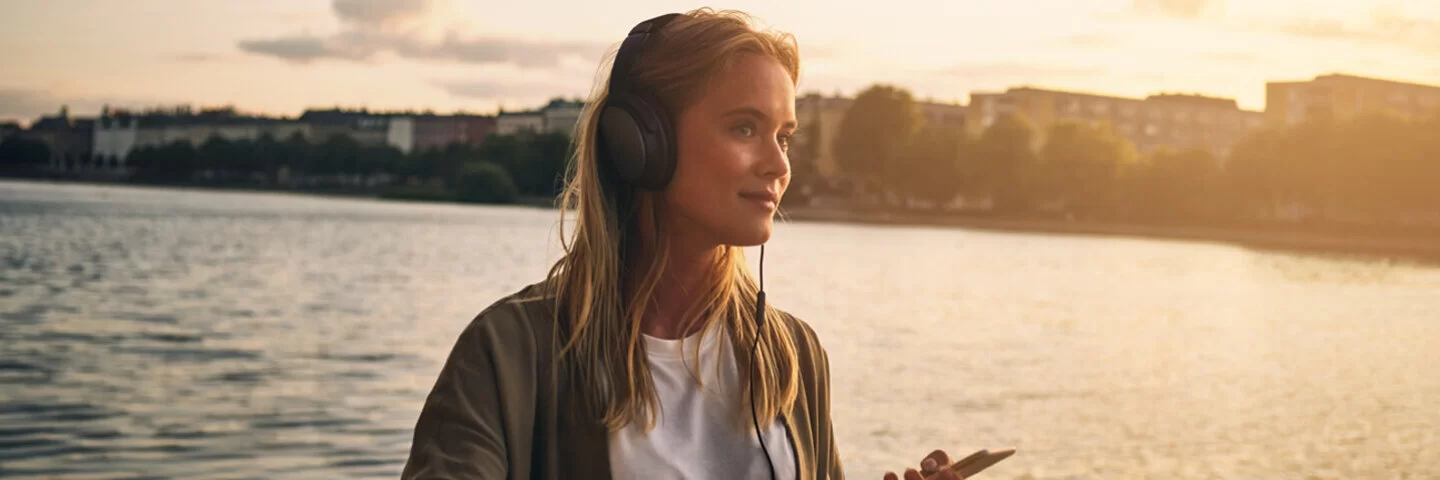 Eine junge Frau steht an einem Flussufer und hört mit Kopfhörern einen Podcast.