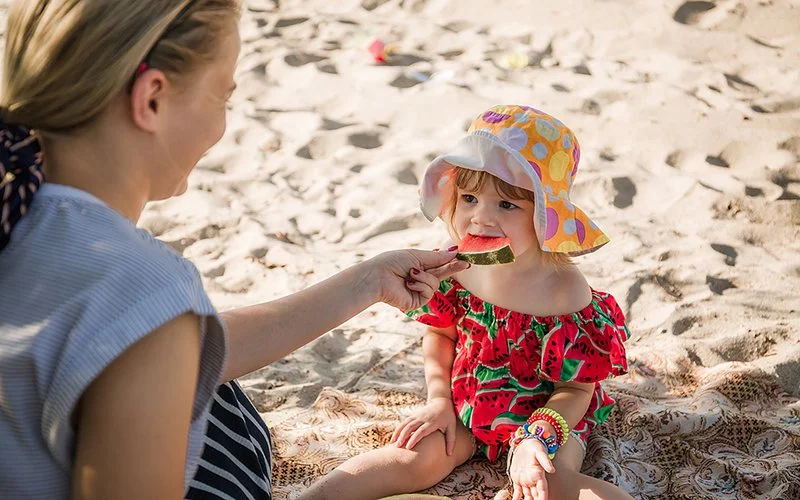 Ein kleines Mädchen sitzt im Schatten auf einer Decke am Strand. Sie trägt einen Sonnenhut und beißt in ein Stück Wassermelone, das die Mutter ihr reicht.