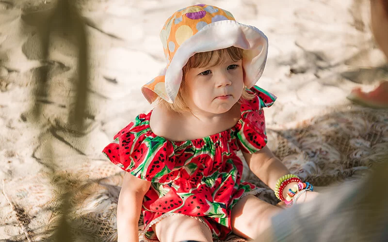 Ein kleines Mädchen sitzt im Schatten auf einer Decke am Strand. Sie trägt einen Sonnenhut und beißt in ein Stück Wassermelone, das die Mutter ihr reicht.