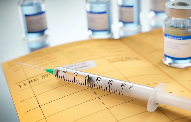 Auf einem aufgeschlagenen Impfpass liegt eine Spritze und ein Fläschchen mit Impfstoff steht darauf. Das Impfdatum ist eingetragen und das internationale Impfzertifikat eingeklebt.