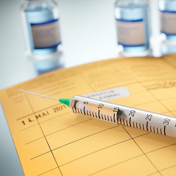 Auf einem aufgeschlagenen Impfpass liegt eine Spritze und ein Fläschchen mit Impfstoff steht darauf. Das Impfdatum ist eingetragen und das internationale Impfzertifikat eingeklebt.