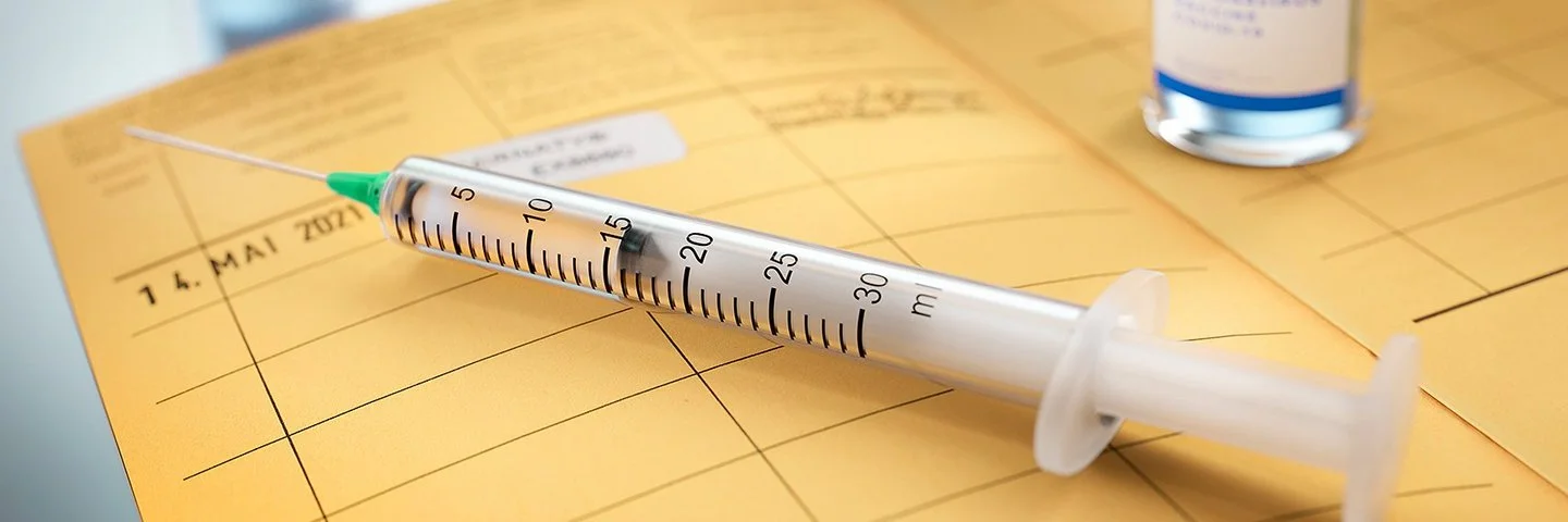 Auf einem aufgeschlagenen Impfpass liegt eine Spritze und ein Fläschchen mit Impfstoff steht darauf. Das Impfdatum ist eingetragen und das internationale Impfzertifikat eingeklebt.