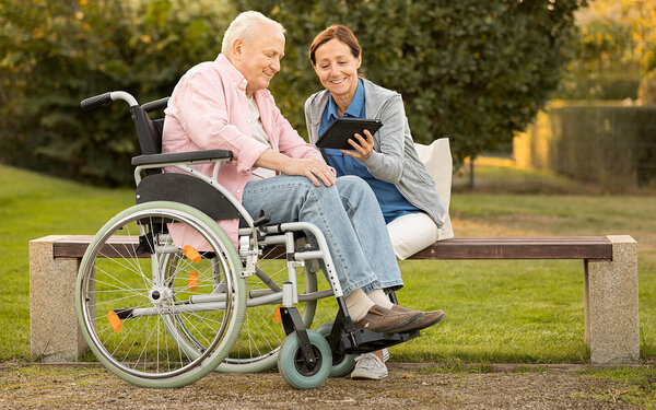 Ein an der Autoimmunerkrankung Multiple Sklerose Erkrankter im Rollstuhl schaut sich zusammen mit seiner Begleiterin im Park etwas auf einem Tablet an.