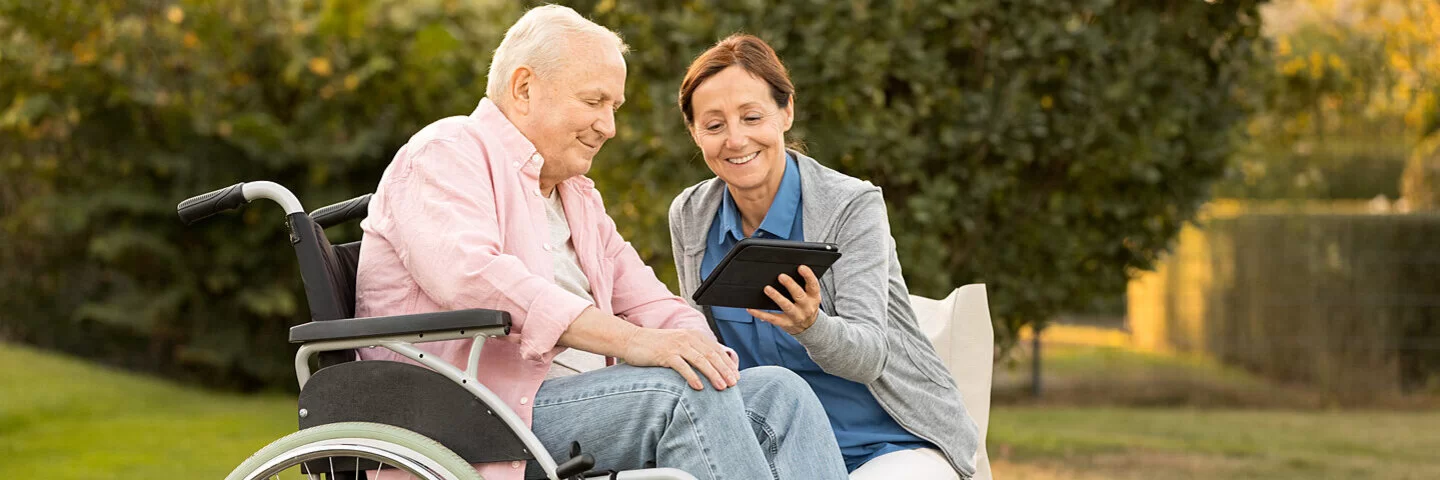 Ein an der Autoimmunerkrankung Multiple Sklerose Erkrankter im Rollstuhl schaut sich zusammen mit seiner Begleiterin im Park etwas auf einem Tablet an.