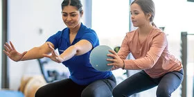 Eine Physiotherapeutin macht mit einem Mädchen Übungen mit einem Ball. Kinder lernen in der Reha, mit Erkrankungen umzugehen.