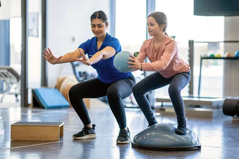 Eine Physiotherapeutin macht mit einem Mädchen Übungen mit einem Ball. Kinder lernen in der Reha, mit Erkrankungen umzugehen.