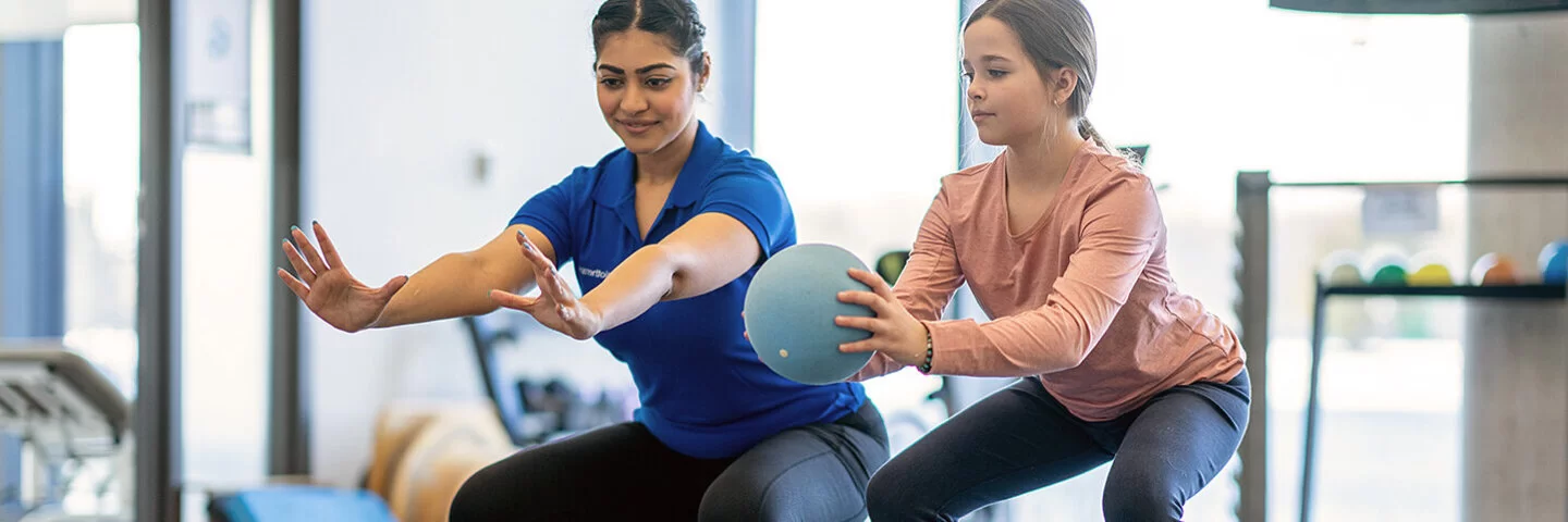 Eine Physiotherapeutin macht mit einem Mädchen Übungen mit einem Ball. Kinder lernen in der Reha, mit Erkrankungen umzugehen.
