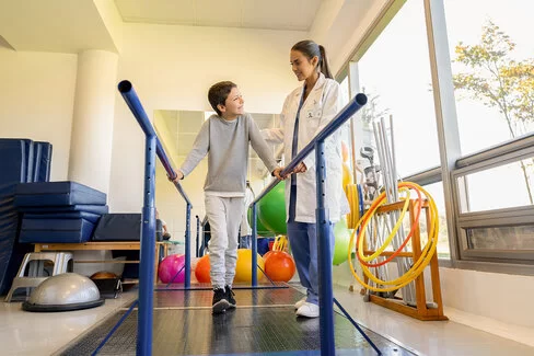 Eine Physiotherapeutin macht mit einem Jungen Gehübungen. Kinder lernen in der Reha, mit Erkrankungen umzugehen.