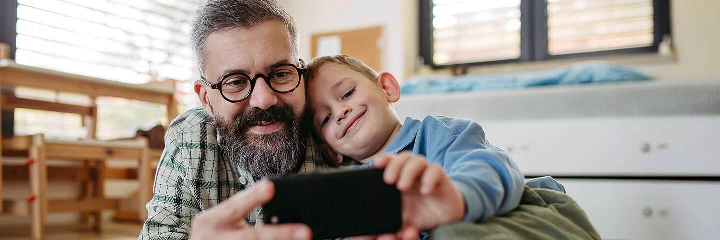 Ein Vater liegt zusammen mit seinem Sohn auf dem Boden im Kinderzimmer. Beide lächeln und schauen auf ein Smartphone, das sie gemeinsam in den Händen halten.