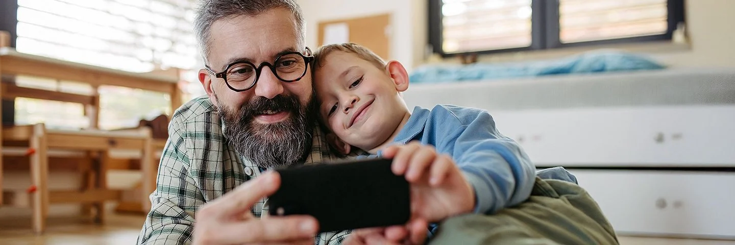 Ein Vater liegt zusammen mit seinem Sohn auf dem Boden im Kinderzimmer. Beide lächeln und schauen auf ein Smartphone, das sie gemeinsam in den Händen halten.