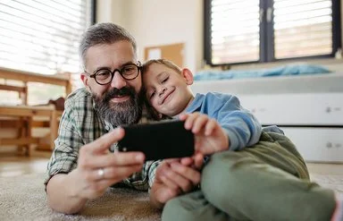 Ein Vater liegt zusammen mit seinem Sohn auf dem Boden im Kinderzimmer. Beide lächeln und schauen auf ein Smartphone, das sie gemeinsam in den Händen halten.