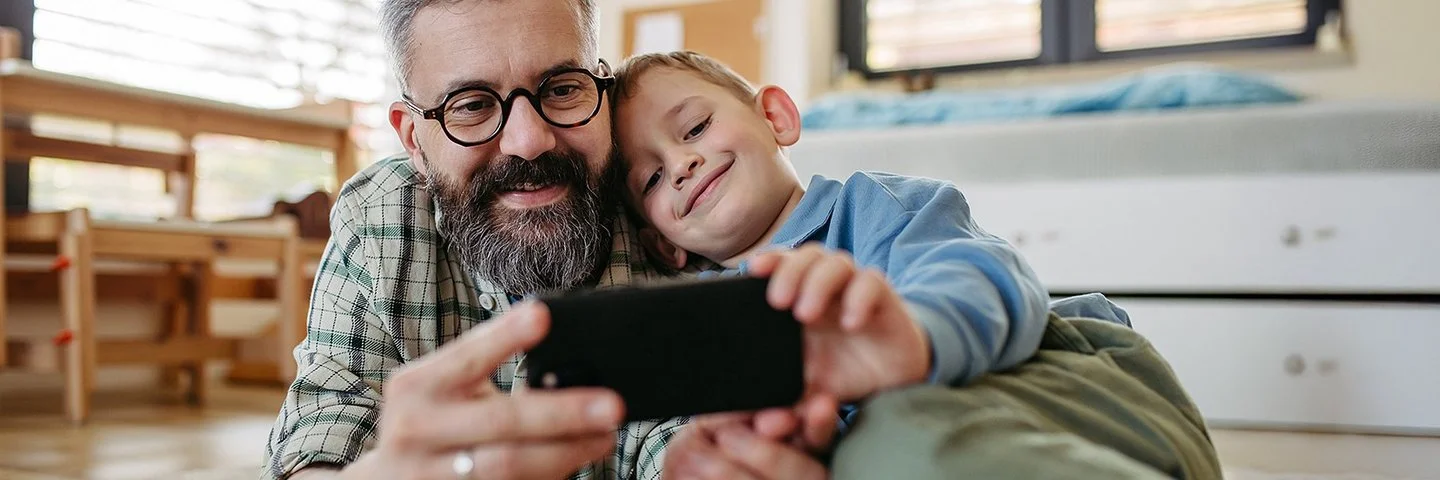 Ein Vater liegt zusammen mit seinem Sohn auf dem Boden im Kinderzimmer. Beide lächeln und schauen auf ein Smartphone, das sie gemeinsam in den Händen halten.