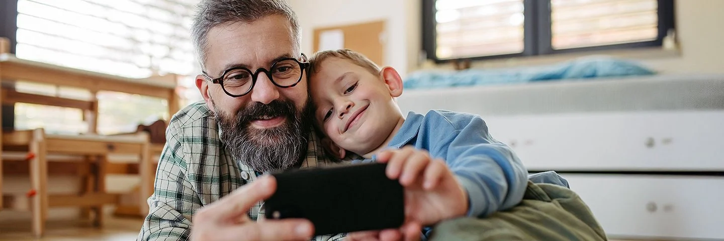 Ein Vater liegt zusammen mit seinem Sohn auf dem Boden im Kinderzimmer. Beide lächeln und schauen auf ein Smartphone, das sie gemeinsam in den Händen halten.