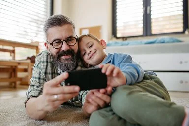 Ein Vater liegt zusammen mit seinem Sohn auf dem Boden im Kinderzimmer. Beide lächeln und schauen auf ein Smartphone, das sie gemeinsam in den Händen halten.