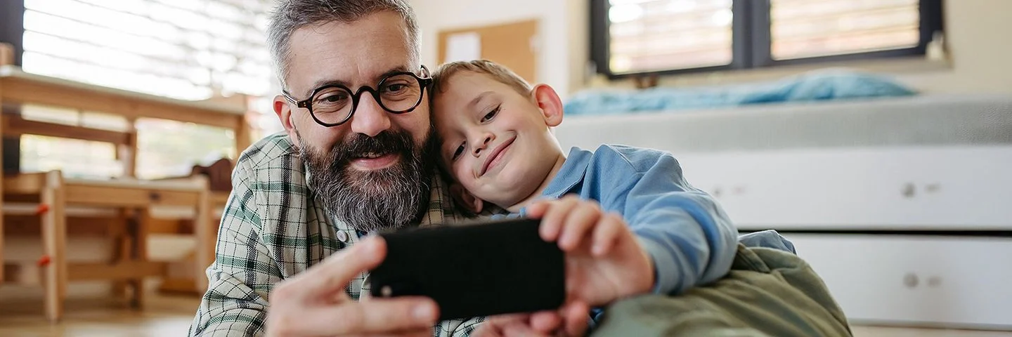 Ein Vater liegt zusammen mit seinem Sohn auf dem Boden im Kinderzimmer. Beide lächeln und schauen auf ein Smartphone, das sie gemeinsam in den Händen halten.